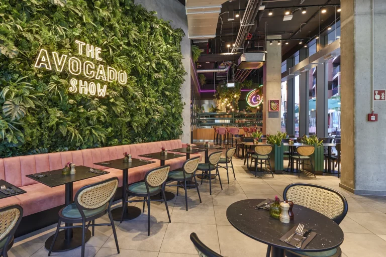 Die begrünte Wand mit dem leuchtenden Logo „The Avocado Show“ und einem Neon-Avocado-Schild im hinteren Bereich prägt das Ambiente. Davor verläuft eine lange Sitzbank mit rosa Polster, ergänzt durch schwarze Holzstühle mit Geflecht-Rückenlehnen und grünen Sitzpolstern. Schwarze Tische in Marmoroptik, Pflanzeninseln und farbige Lichtakzente strukturieren den Raum und schaffen eine moderne, frische Atmosphäre.